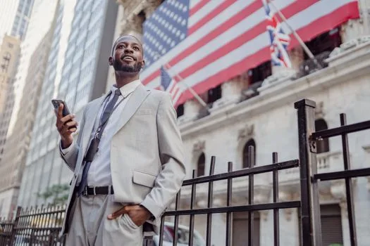 Confident man in a gray suit holding a cellphone outside a building with flags, signifying business success.